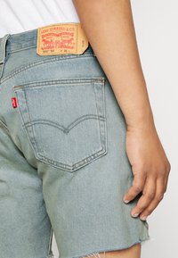 Person wearing light blue denim Levi's shorts with back pocket and leather brand patch, hand relaxed by side against white background.