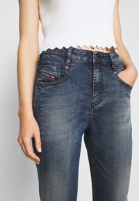 Vaqueros de mezclilla en un lavado oscuro con una textura ligeramente desgastada, que presentan un pequeño bolsillo decorativo, presillas para cinturón y cierre frontal con botón de metal.