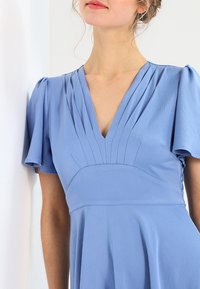 Lichtblauwe jurk met geplooide V-hals, korte mouwen en een aansluitend lijfje dat uitloopt in een uitlopende rok. Gladde, gestructureerde stof.