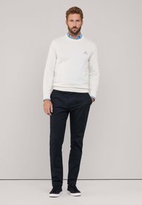 Maglione bianco, colletto blu della camicia, pantaloni scuri, sneakers nere. Design semplice, texture liscia, dettaglio del logo sul davanti. Vista a lunghezza intera.