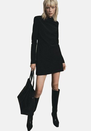 Vestido mini negro de mangas largas con cuello alto, combinado con botas negras hasta la rodilla y un bolso tote negro a juego. Textura suave.