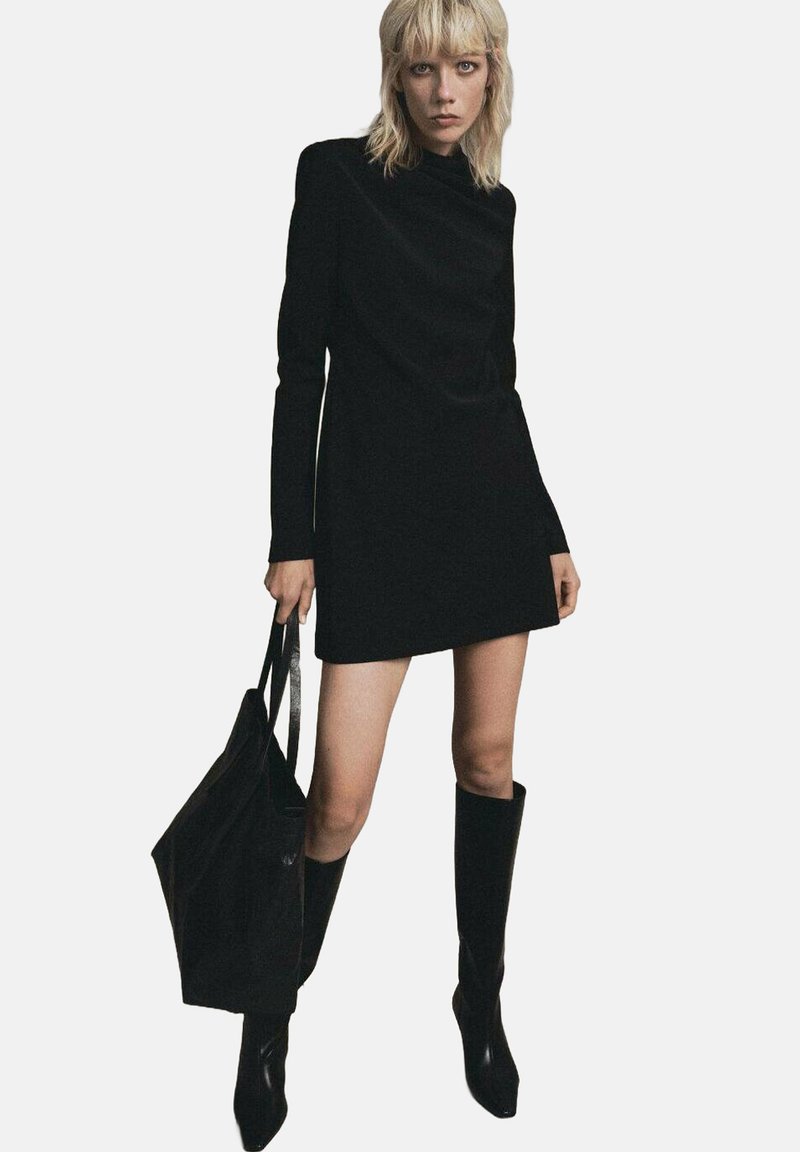 Vestido mini negro de mangas largas con cuello alto, combinado con botas negras hasta la rodilla y un bolso tote negro a juego. Textura suave.