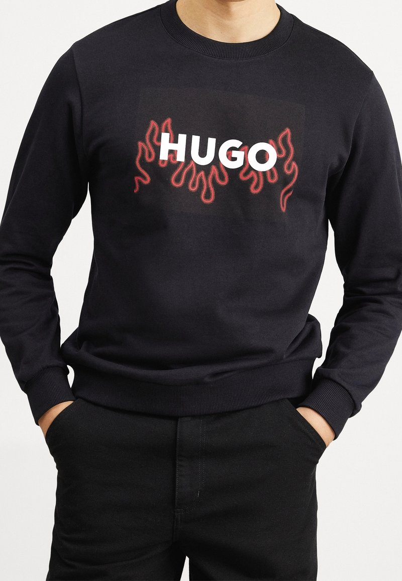 Sweat-shirt noir avec motifs de flammes rouges et texte blanc en gras « HUGO » sur la poitrine, porté avec un pantalon noir assorti. Mains dans les poches.