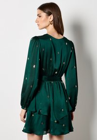 Groene satijnen jurk met lange mouwen, gerimpelde lagen en gouden bladerenversieringen. Heeft een nauwsluitende taille en een ronde halslijn.