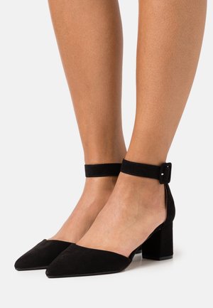 Sorte ruskinds høje hæle med spids tå, ankelsstrap sikret med en spænde og en blokhæl. Glat tekstur, minimalistisk design.