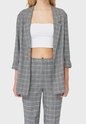 Woman wearing gray plaid blazer and matching pants with white strapless crop top, standing against plain background.