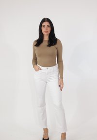 Kobieta o długich ciemnych włosach, ubrana w beżową bluzkę z długim rękawem, białe dżinsy i czarne szpilki, pozująca na jednolitym białym tle.