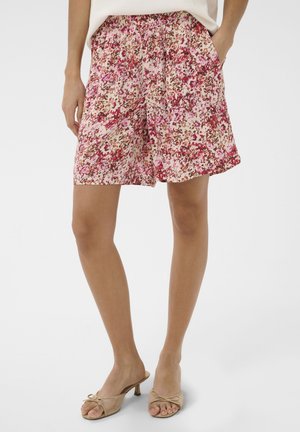 Kobieta w różowych, czerwonych i beżowych szortach w abstrakcyjny kwiatowy wzór z elastyczną talią oraz beżowych sandałach na niskim obcasie z odkrytymi palcami.