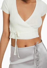 Vit ribbade crop top med djup V-ringning, korta ärmar och en knytning i midjan. Matchad med en silver satin kjol med snörningsdetaljer.