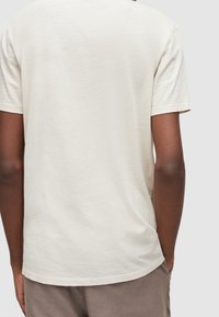 T-shirt en coton beige avec une coupe décontractée, un col rond et des manches courtes. Le tissu a une apparence légèrement texturée et légère.