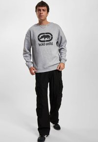 Gray sweatshirt with a black graphic logo featuring an animal and text. Paired with loose-fitting black cargo pants and dark sneakers.