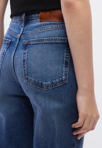 Μαντηλάκια από σκούρο μπλε τζιν με ψηλή μέση, με πέντε τσέπες και ένα καφέ δερμάτινο λογότυπο  στο πίσω μέρος της μέσης.