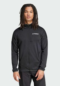 Jacket preta Terrex com fecho, gola alta, mangas longas e bolso no peito. Apresenta uma textura suave e um logótipo branco do lado esquerdo.