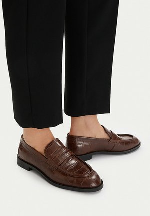 Brązowe mokasyny ze skóry o fakturze krokodyla noszone na bose stopy z czarnymi spodniami o skróconej długości.