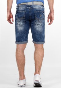 Denim shorts in medium blue met opgerolde manchetten, voorzien van twee achterzakken met stikdetails en een versleten uitstraling. Witte sneakers zichtbaar.