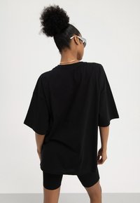 Woman with curly hair in a bun wears oversized black t-shirt, black cycling shorts, silver necklace, multiple ear cuffs, and white futuristic sunglasses.