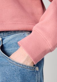 Felpa rosa chiaro con polsini a coste, abbinata a jeans in denim blu strappati. Una mano è appoggiata sulla tasca dei jeans.