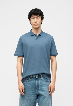 Fiatal férfi kék rövid ujjú galléros pólót és világoskék farmert visel, egyszerű fehér háttér előtt áll.