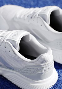 Paire de chaussures de sport blanches avec semelles texturées sur une surface bleue, montrant les détails latéraux et arrière avec les lacets défaits.