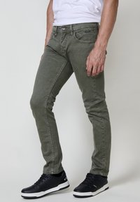 Olivno zelene skinny jeans iz denima, z značilnim pet-pocket dizajnom in subtilnimi detajli šivanja, v kombinaciji s črnimi supergami.