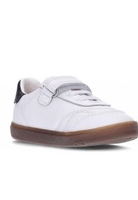 Zapatilla baja blanca con suela marrón, tabique negro en el talón y una sola tira de velcro sobre los cordones blancos.