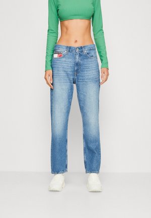 Person iført højtaljede blå jeans, hvide sneakers og en grøn langærmet cropped top, som står mod en ensfarvet hvid baggrund.