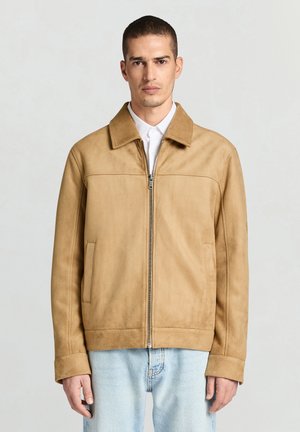 Veste en suède marron clair avec col ouvert, fermeture éclair à l'avant et poches latérales. Présente une texture lisse et une coupe décontractée, associée à un jean bleu clair.