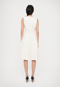 Λευκό αμάνικο φόρεμα με εφαρμοστό μπούστο και φαρδιά φούστα. Διαθέτει φερμουάρ στο πίσω μέρος και ομαλή υφή. Φοράται με μαύρα ψηλοτάκουνα παπούτσια.