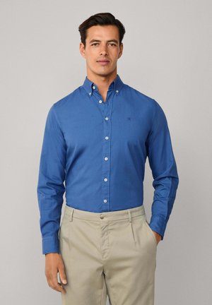 Camicia blu con colletto, realizzata in tessuto liscio, con bottoni bianchi e un logo ricamato discreto. Abbinata a pantaloni leggeri color khaki.