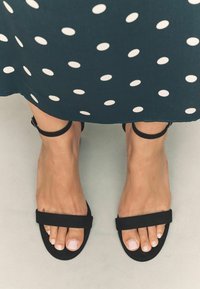 Sandales plates noires avec deux larges sangles, mettant en avant un matériau texturé ; les ongles sont soigneusement manicurés. L'arrière-plan présente un motif à pois.