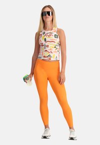 Top corto sportivo con motivi colorati di montagne e alberi, abbinato a leggings arancioni a vita alta e scarpe da ginnastica bianche.