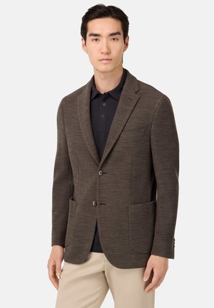 Brauner strukturierter Blazer mit reversähnlichen Kragen, zwei Fronttaschen und zwei Knöpfen; kombiniert mit einem dunklen Poloshirt und beigen Hosen.