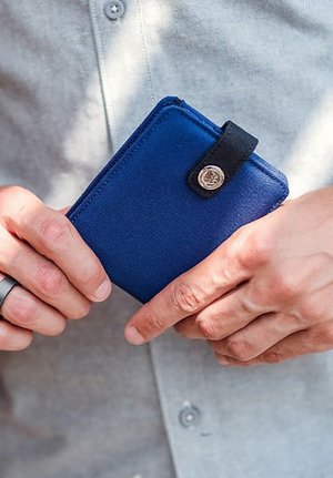 Blue fabric wallet with a black strap and silver snap closure, held in hands. The wallet features a rectangular shape and smooth texture.