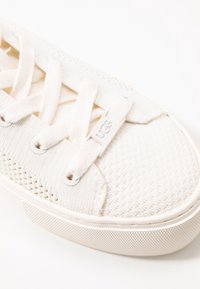 Białe dziane trampki z grubą gumową podeszwą, płaskimi sznurówkami i plastikowym znaczkiem na sznurówce z wytłoczonym logo "UGG", widok pod kątem na strefę palców.