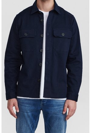 Homme portant une veste-chemise bleu marine boutonnée sur un t-shirt blanc et un jean bleu, debout, les mains détendues le long du corps.