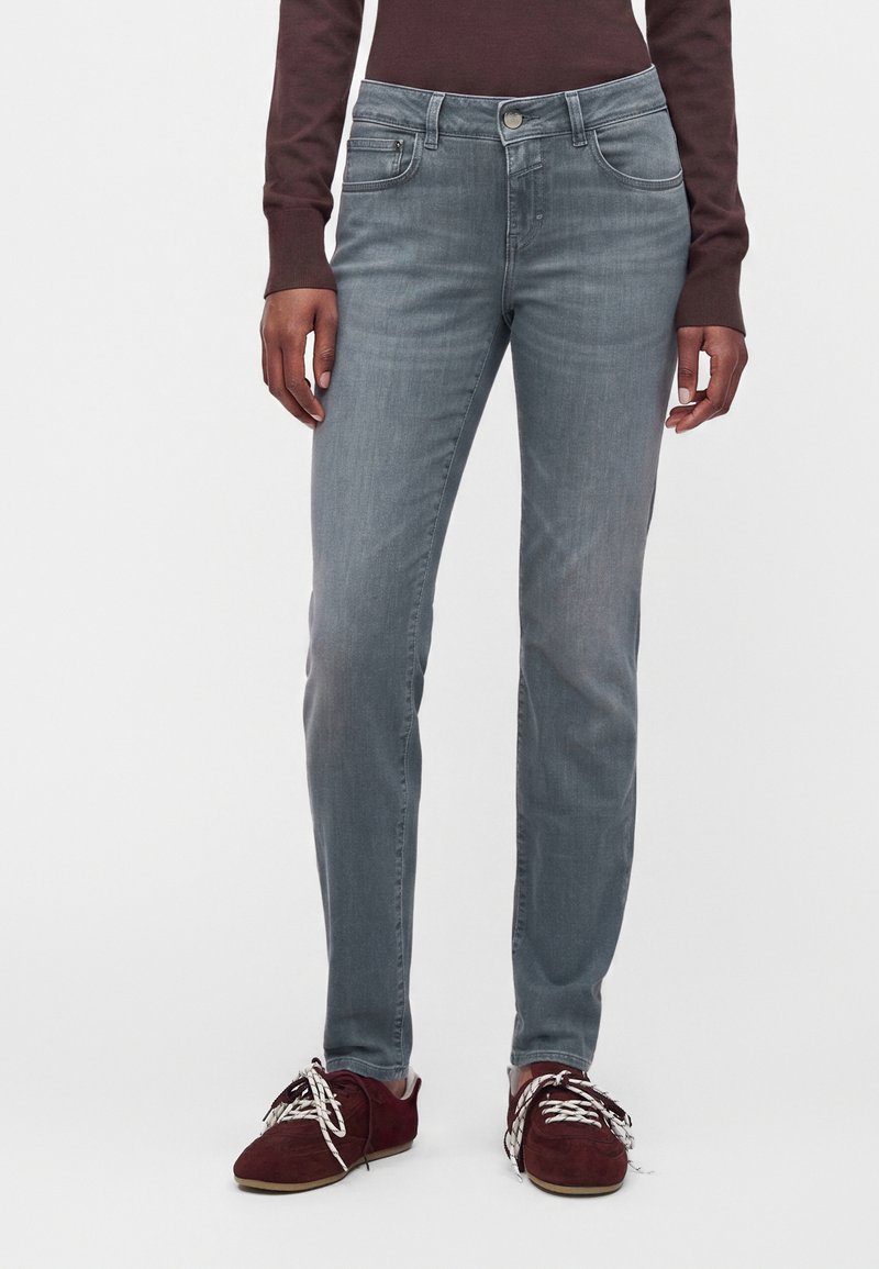 Personne portant un jean gris ajusté, un haut à manches longues bordeaux foncé et des chaussures bordeaux avec des lacets blancs, debout devant un fond blanc uni.