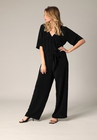 Combinaison noire à manches courtes, décolleté en V et taille cintrée. Le tissu a un léger éclat. Associée à des sandales noires à talons hauts.