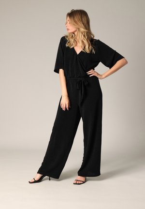 Zwarte jumpsuit met korte mouwen, een V-halslijn en een strikomhoog. De stof heeft een subtiele glans. Gecombineerd met zwarte sandalen met hoge hakken.