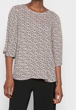 Femme portant un chemisier blanc à imprimé floral noir miniature et manches trois-quarts, associé à un pantalon noir.