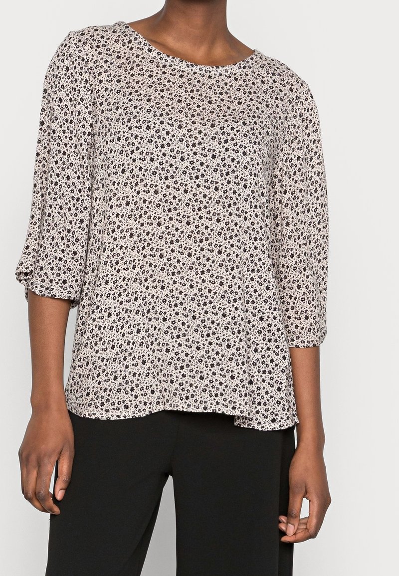 Femme portant un chemisier blanc à imprimé floral noir miniature et manches trois-quarts, associé à un pantalon noir.