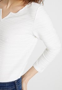Maglietta bianca a maniche lunghe con un design a coste testurizzato e scollatura a V. Abbinata a jeans blu scuro.