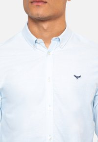 Camisa de botones azul claro con cuello, que presenta un pequeño logo bordado en navy en el lado izquierdo del pecho y botones blancos para el cierre.