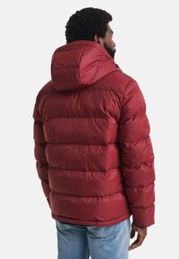 Mann in einer weinrot gefüllten Jacke mit Kapuze, von hinten betrachtet, kombiniert mit hellblauen Jeans vor einem weißen Hintergrund.