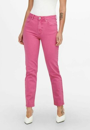 Persoon met een roze high-waisted jeans, wit mouwloos topje, witte hakken; tatoeage zichtbaar op de rechter onderarm en de linker enkel; effen achtergrond.