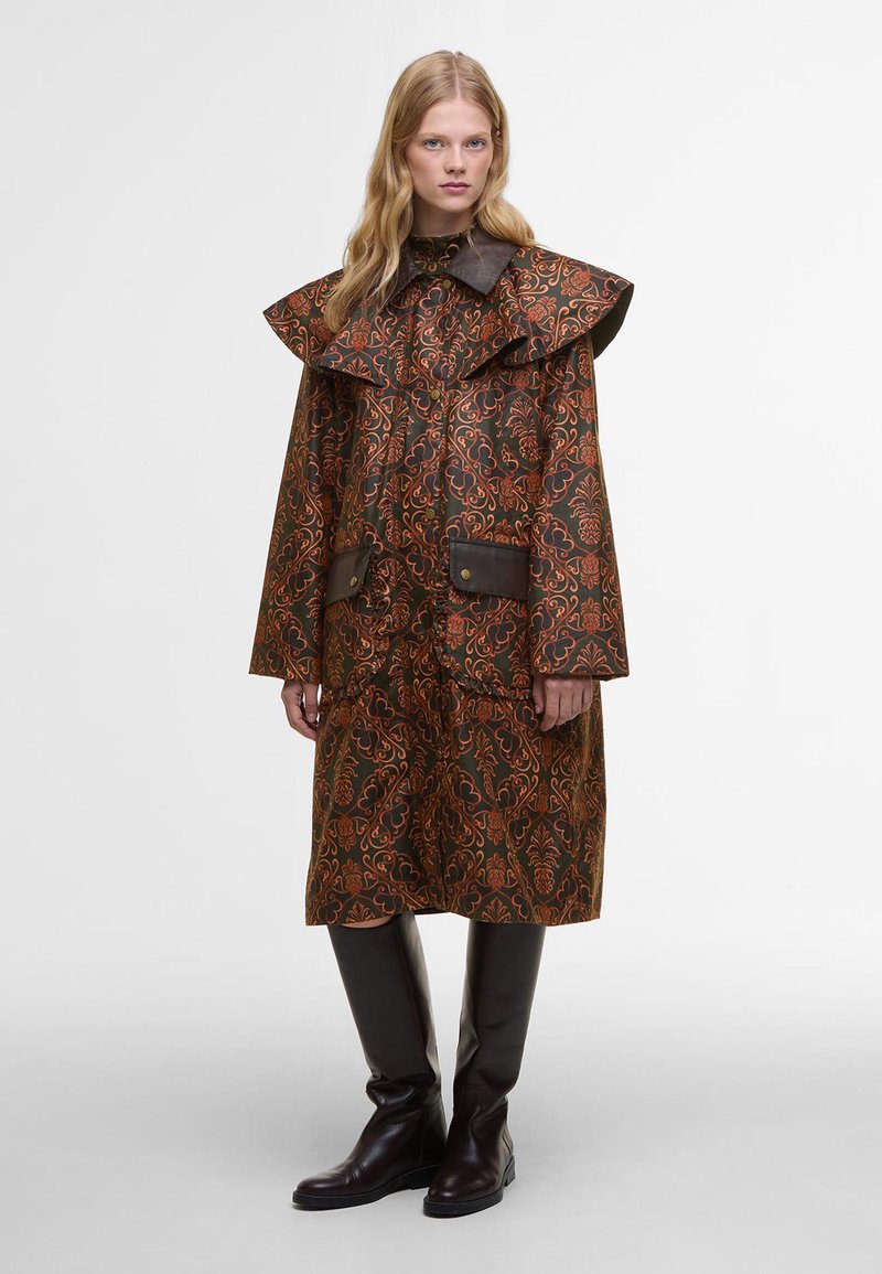 Brauner gemusterter Mantel mit großem, rüschenbesetztem Kragen, der dunkle braune Flecken und Knöpfe aufweist, getragen über figurbetonten, knielangen braunen Stiefeln.