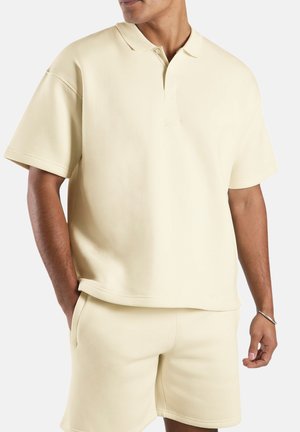 Hombre con camisa polo de manga corta color crema y pantalones cortos a juego, con pulsera en la muñeca derecha, manos en los bolsillos, sobre fondo liso.