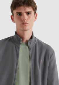 Grijze zip-up jack met een textuurdesign, hoge kraag en subtiele logo-accent. Draagt over een lichtgroen T-shirt.
