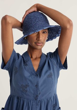 Kobieta w niebieskiej sukience z haftem i guzikami oraz niebieskim szerokim kapeluszu z plecionki, pozująca z uniesionymi rękami za głową.