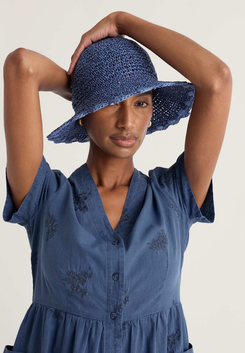 Mujer con un vestido azul bordado y abotonado, y un sombrero azul de ala ancha tejido, posando con los brazos levantados detrás de la cabeza.