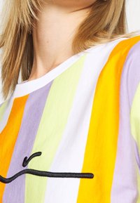 Henkilö, joka on pukeutunut moniväriseen raidalliseen paitaan, jossa on pystysuoria oranssin, violetin, keltaisen ja vihreän väriä sekä musta brodeerattu viivakuvaus.